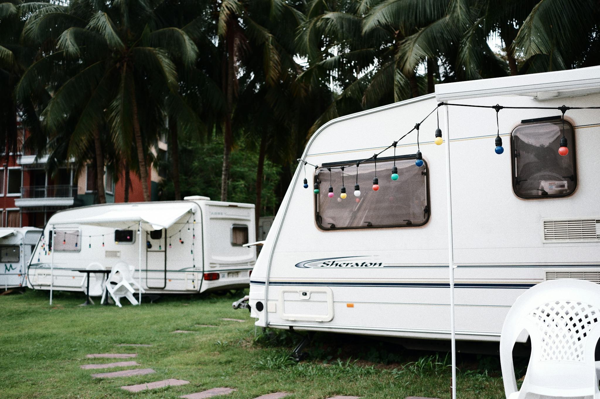 Relaxing caravan camping site under palm trees with outdoor seating and festive lights.